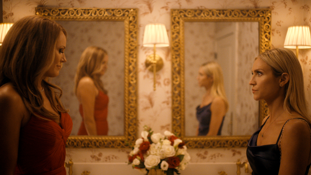 "Two women in elegant dresses stand facing each other in a warmly lit, ornate bathroom with floral wallpaper, gold-framed mirrors, wall lamps, and a vase of flowers on the counter, their reflections visible in the mirrors."