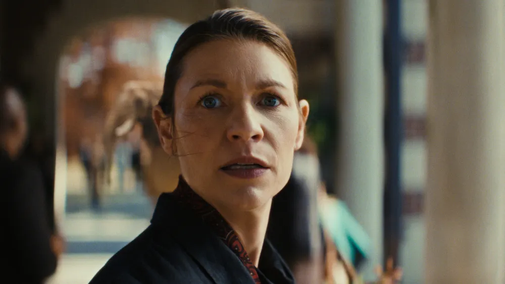 "A woman with a concerned expression stands outdoors in a sunlit urban setting, surrounded by blurred figures and architectural columns, creating a dramatic and tense mood."