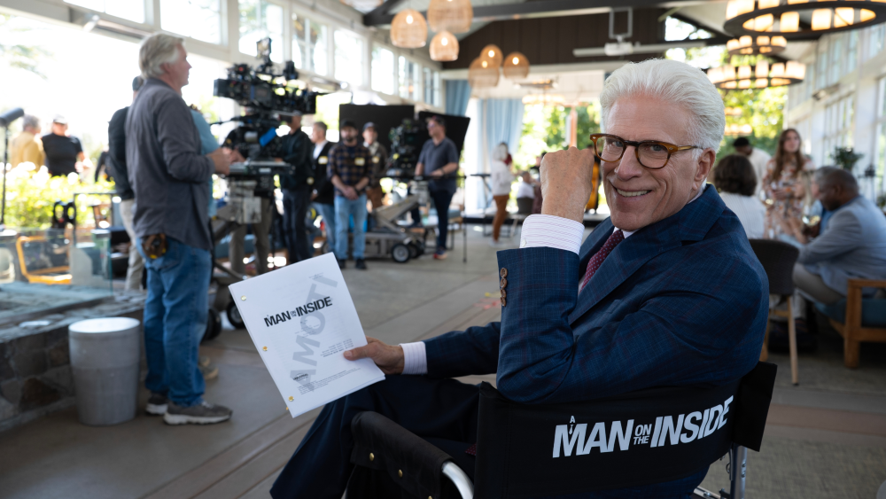 "Man with white hair and glasses sitting in a director’s chair holding a script on a film set, with crew, cameras, and lighting equipment visible in a bright indoor setting."