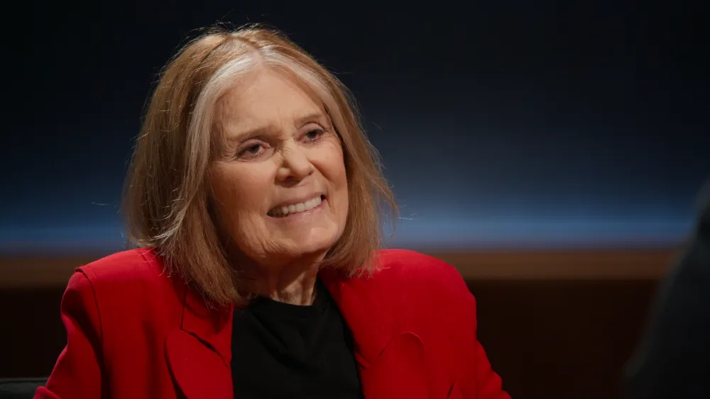 "Smiling elderly woman with shoulder-length light hair, wearing a red blazer and black shirt, sitting indoors in a dimly lit interview or studio setting with a dark, softly illuminated background."
