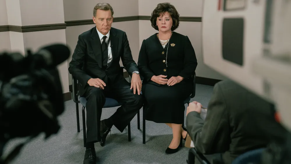 "Two people in formal attire sit side by side in a plain office-like room, holding hands, being filmed by a video camera during an interview or press conference."