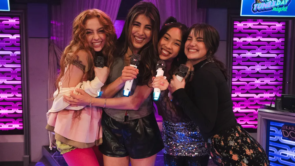 "Four young women smiling and hugging while holding microphones, standing together on a colorful karaoke stage decorated with purple lights and a "Tunes-day Night" sign, suggesting a lively and fun indoor environment."